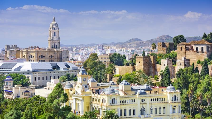 Aerial image overlooking Malaga, Spain's colorful classic buildings with lush trees in the forefront and blue sky in the distance.