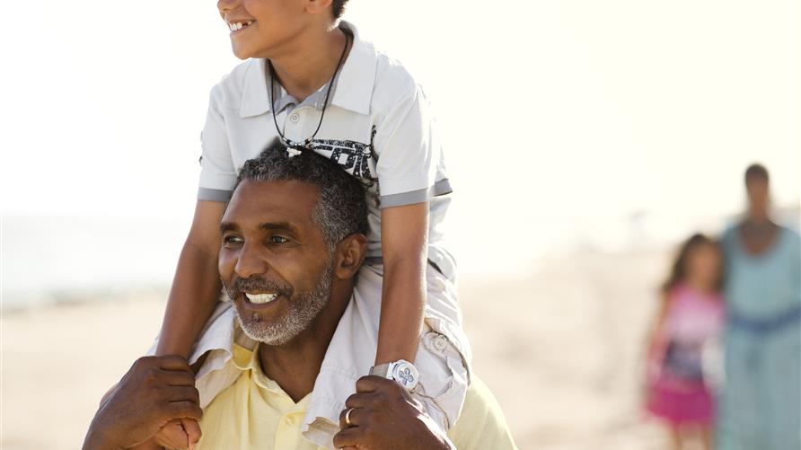 Father, with son on his shoulders, smiling and walking on the beach. 