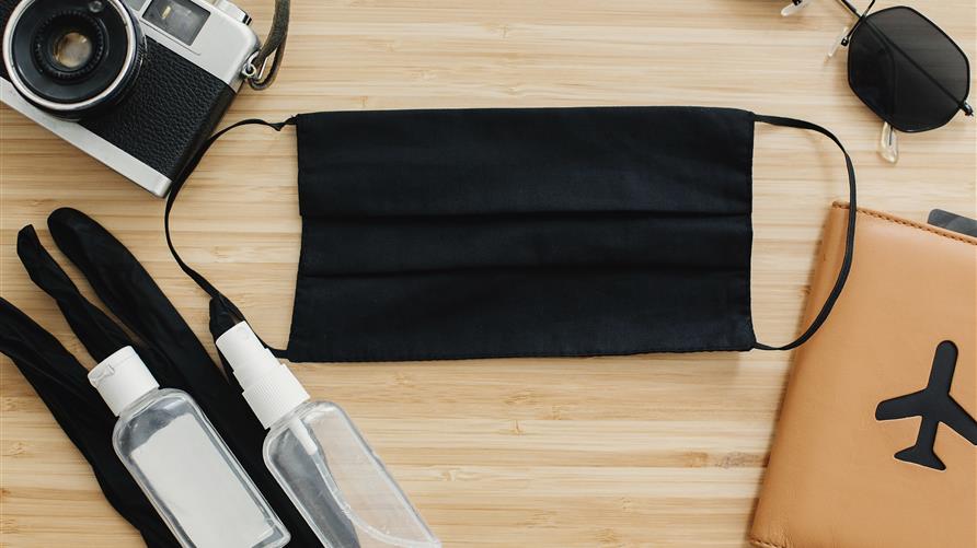 Flat lay shot of COVID-19 travel essentials — face mask, hand sanitizer, camera, and sunglasses. 