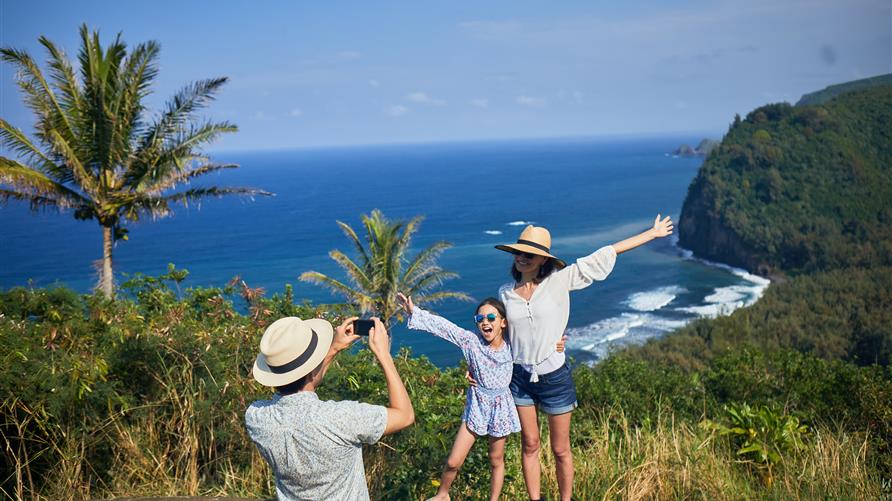 Father taking a picture of mom and daughter happily posing in front of the ocean on vacation in Hawaii. 