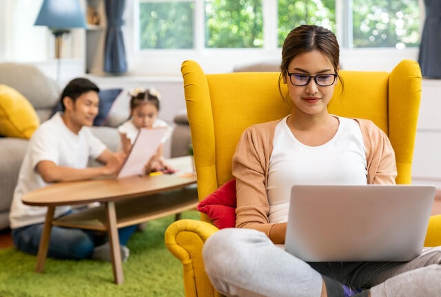 Woman sitting in chair using lap top with young family in the background. 