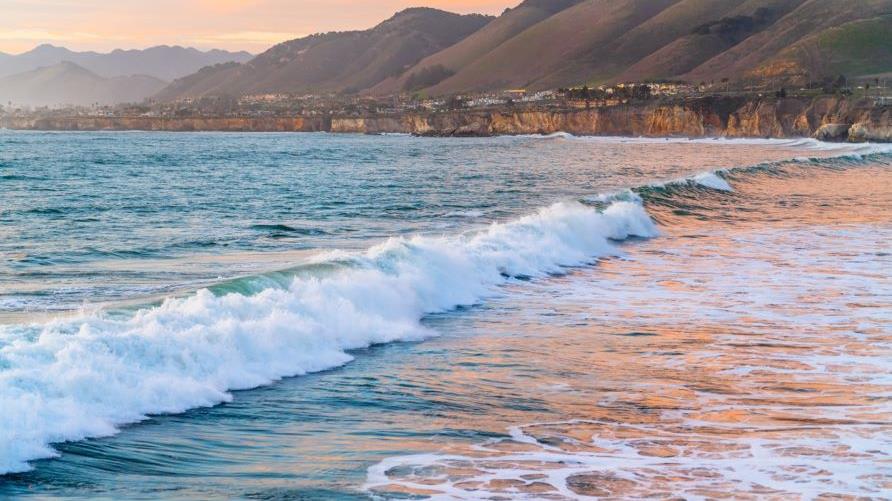 Idyllic coastal scene, waves washing ashore, mountains in the distances, cotton candy colored skies. 