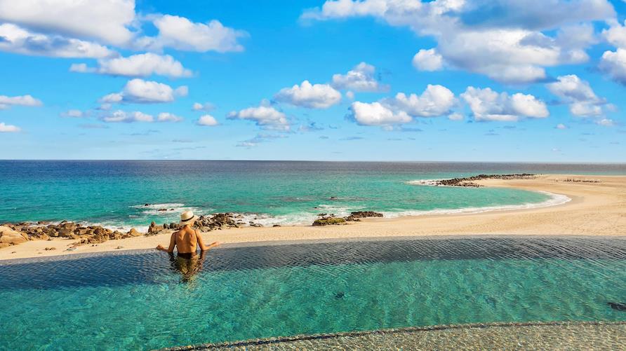 Woman in an infinity pool looks out at a pristine beach and beautiful turquoise water in Los Cabos, Mexico