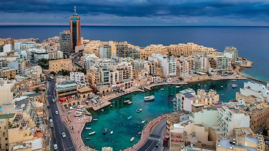 Drone image of Spinola Bay in Malta, featuring emerald waters in the bay, colorful pastel buildings that surround it, and the deep blue ocean just beyond. The skies are cloudy giving a soft glow.