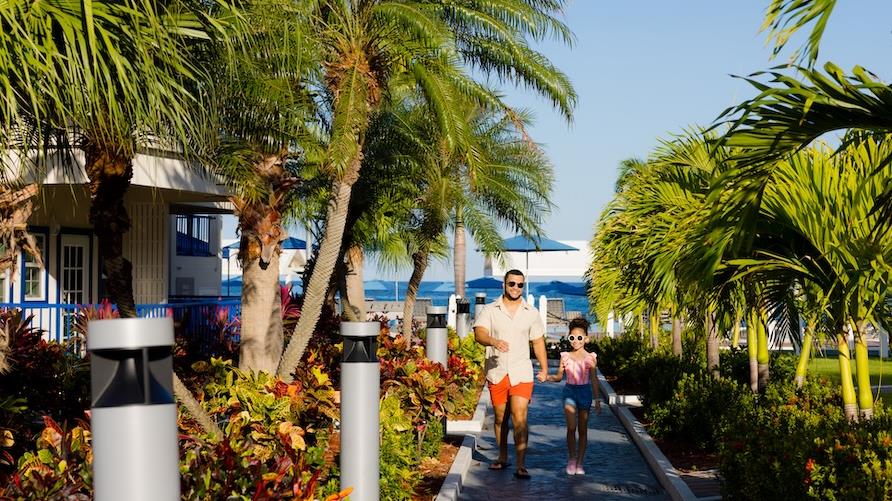 Man and young girl walk under sunny skies along the palm-tree lined path at Flamingo Beach, a Hilton Grand Vacation Club
