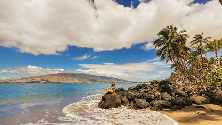 Idyllic beach scene, woman sitting atop rocks, surrounded by crashing waves, golden shoreline, blue skies, palm trees, Maui, Hawaii. 