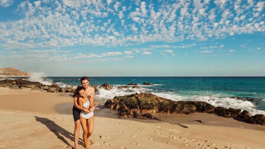 Couple embracing each other happily, beach, waves crashing behind, golden sands, blue skies overhead, Los Cabos, Mexico.