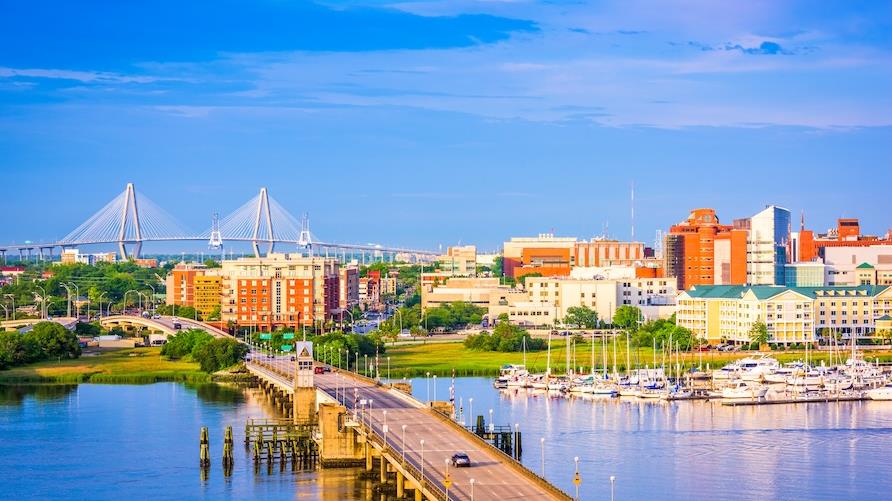 Bright daytime view of the city of Charleston, SC with the bridge and river in the foreground, and the pastel-colored buildings in the background under bright blue skies.
