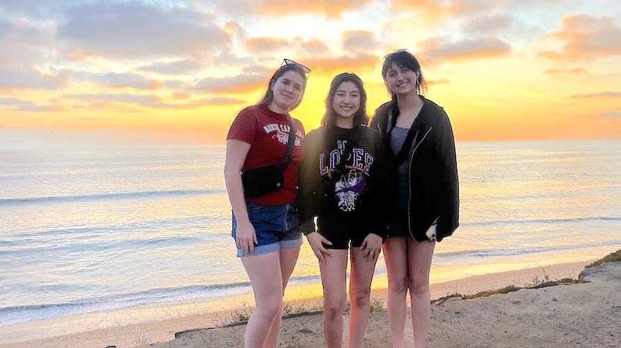 Three young women standing on the shoreline facing the camera against a setting sun in Carlsbad, California
