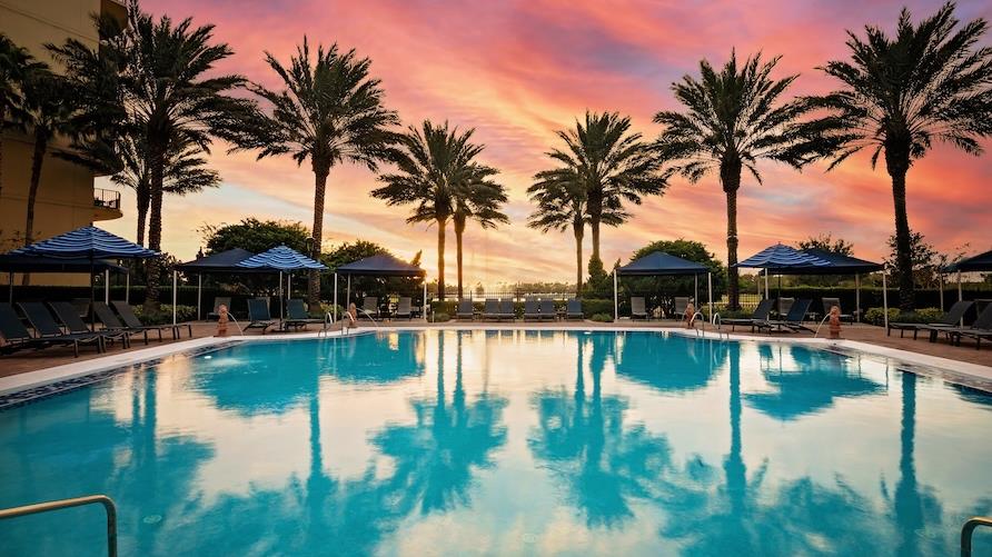 Image of the blue pool at Parc Soleil, a Hilton Grand Vacation Club with a line of palms in the distance and pink and purple hues of a sunset beyond.