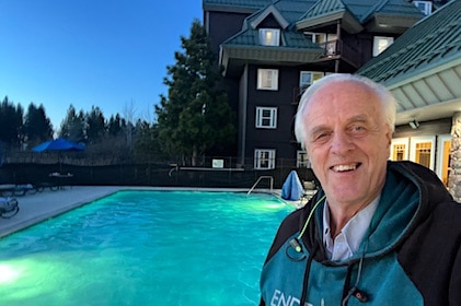 Hilton Grand Vacations Member, Cliff, in a close-up pose in front of lighted pool with pine trees, the resort and blue sky in the background.