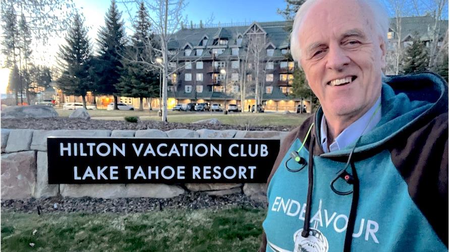 Hilton Grand Vacations Member Cliff B stands in front of the Hilton Vacation Club Lake Tahoe resort sign and stone wall. Pine trees and a long shot of the resort building against a blue sky is in the background.