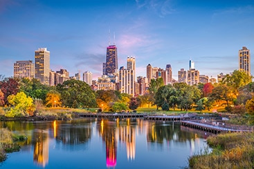 A lake surrounded by trees in autumn colors with tall skyscrapers in the background.