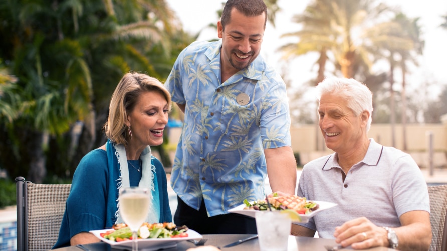 Older couple being served a meal outdoors by a handsome man, surrounded by palm trees