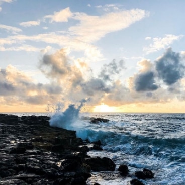 Foamy waves crash onto dark rocks on the Hawaiian beach at sunset.