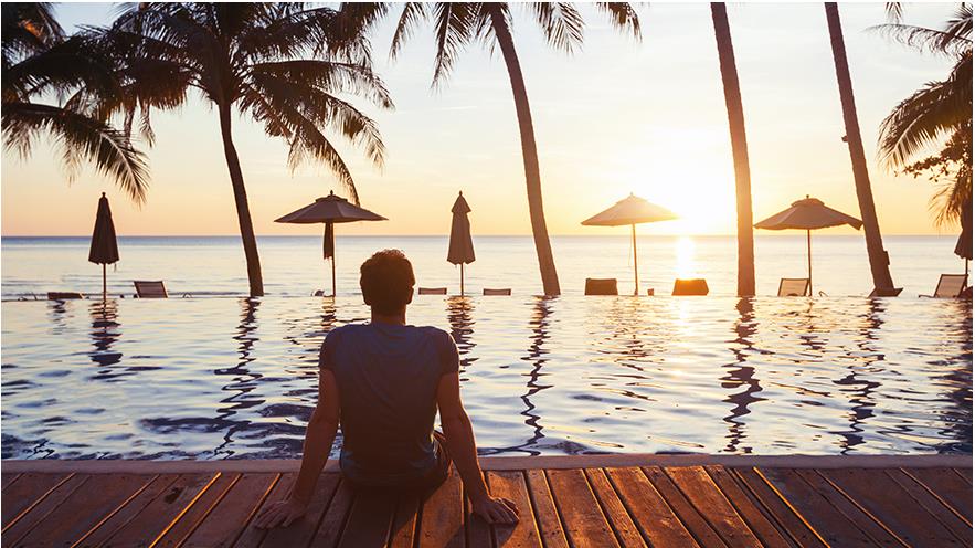 Silhouette of a man sitting on the edge of an infinity pool lined with palm trees overlooking the ocean. 